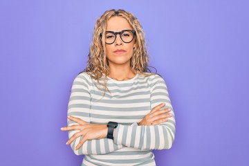 Beautiful blonde woman wearing casual striped t-shirt and glasses over purple background skeptic...