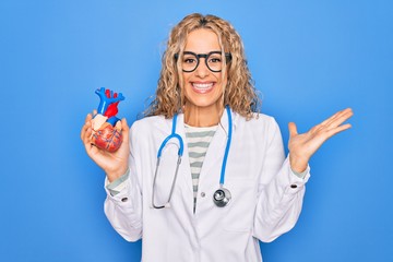 Young beautiful blonde cardiologist woman wearing stethoscope holding plastic heart celebrating victory with happy smile and winner expression with raised hands