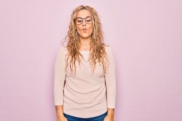 Obraz premium Young beautiful blonde woman wearing casual sweater and glasses over pink background making fish face with lips, crazy and comical gesture. Funny expression.