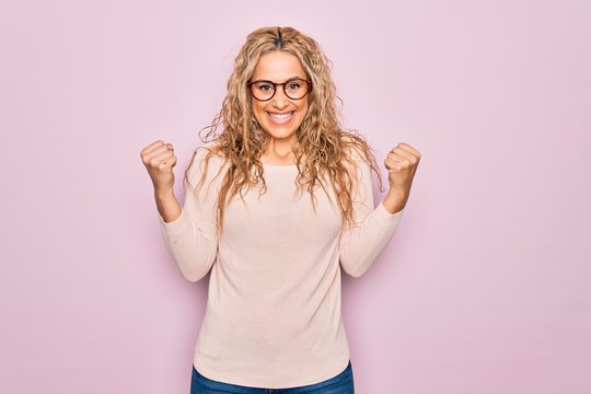 Young beautiful blonde woman wearing casual sweater and glasses over pink background celebrating surprised and amazed for success with arms raised and open eyes. Winner concept.