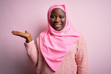 Young african american plus size woman wearing muslim hijab over isolated pink background smiling cheerful presenting and pointing with palm of hand looking at the camera.