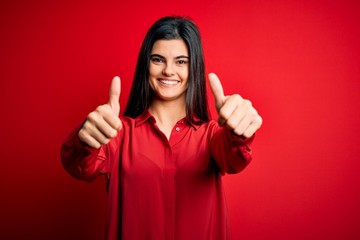 Young beautiful brunette woman wearing casual shirt standing over red background approving doing positive gesture with hand, thumbs up smiling and happy for success. Winner gesture.