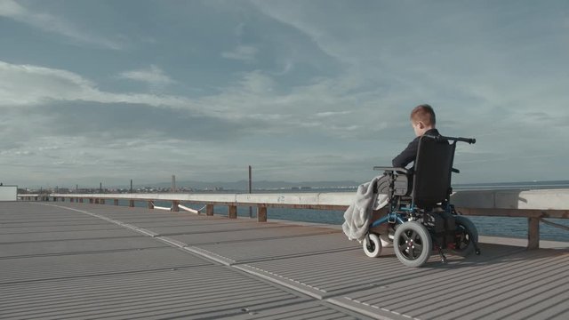 Wide shot of following the child in electric wheelchair. Handicapped boy enjoying fresh air during the ride along seafront