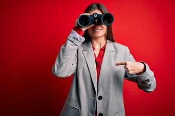 Young beautiful woman with blue eyes using binoculars over isolated red background with surprise face pointing finger to himself