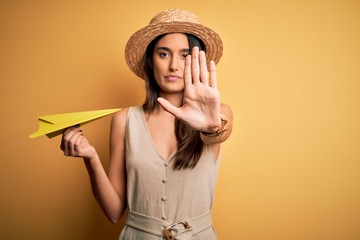 Young beautiful brunette woman wearing hat holding paper airplane over yellow background with open hand doing stop sign with serious and confident expression, defense gesture