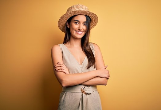Young beautiful brunette woman on vacation wearing casual dress and hat happy face smiling with crossed arms looking at the camera. Positive person.