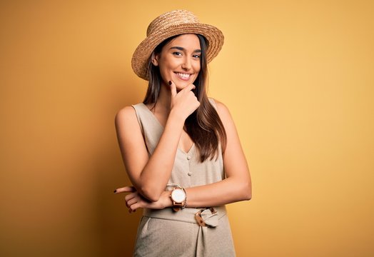 Young beautiful brunette woman on vacation wearing casual dress and hat looking confident at the camera smiling with crossed arms and hand raised on chin. Thinking positive.