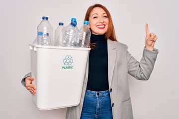 Young beautiful redhead woman recycling holding trash can with plastic bottles to recycle surprised...