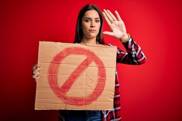 Young beautiful brunette woman holding banner with prohibited signal over red background with open...