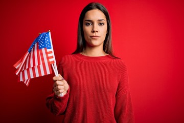 Young beautiful brunette patriotic woman holding united states flag over red background with a confident expression on smart face thinking serious