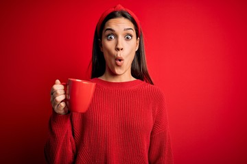 Young beautiful brunette woman drinking mug of coffe over isolated red background scared in shock with a surprise face, afraid and excited with fear expression
