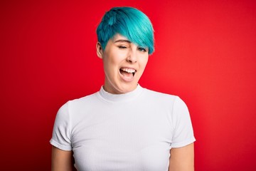 Young beautiful woman with blue fashion hair wearing casual t-shirt over red background winking...