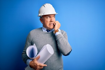 Middle age handsome grey-haired architect man wearing safety helmet holding blueprints looking...