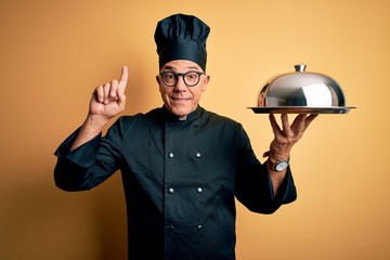 Middle age handsome grey-haired waiter man wearing cooker uniform and hat holding tray smiling...