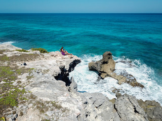 A beautiful young woman sitting  at the cliff rock looking into the deep turquoise waters of the Caribbean ocean, shot on  a drone, view from the bird eye 