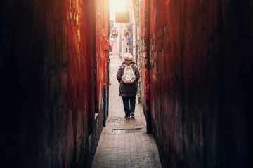 Traveler girl leaves narrow long red corridor or tunnel on city street, rear view.