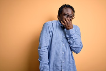 Young handsome african american man wearing shirt and glasses over yellow background smelling...