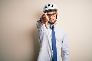 Young businessman wearing glasses and bike helmet standing over isolated white bakground looking unhappy and angry showing rejection and negative with thumbs down gesture. Bad expression.