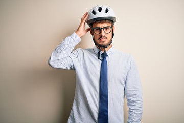 Young businessman wearing glasses and bike helmet standing over isolated white bakground confuse and wonder about question. Uncertain with doubt, thinking with hand on head. Pensive concept.