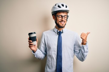 Young businessman wearing glasses and bike helmet drinking cup of coffee pointing and showing with thumb up to the side with happy face smiling