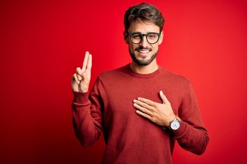 Young handsome man with beard wearing glasses and sweater standing over red background smiling...