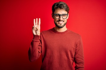 Young handsome man with beard wearing glasses and sweater standing over red background showing and pointing up with fingers number three while smiling confident and happy.