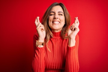 Young beautiful brunette woman wearing casual turtleneck sweater over red background gesturing finger crossed smiling with hope and eyes closed. Luck and superstitious concept.