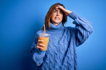 Young beautiful brunette woman drinking healthy orange juice over blue background stressed with hand on head, shocked with shame and surprise face, angry and frustrated. Fear and upset for mistake.