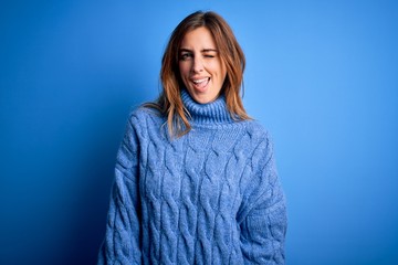 Young beautiful brunette woman wearing casual turtleneck sweater over blue background winking...