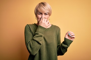 Young blonde woman with modern short hair wearing casual sweater over yellow background smelling...