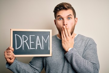 Young blond man with beard and blue eyes holding blackboard with dream message cover mouth with hand shocked with shame for mistake, expression of fear, scared in silence, secret concept