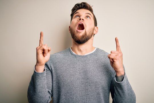 Young handsome blond man with beard and blue eyes wearing casual sweater amazed and surprised looking up and pointing with fingers and raised arms.