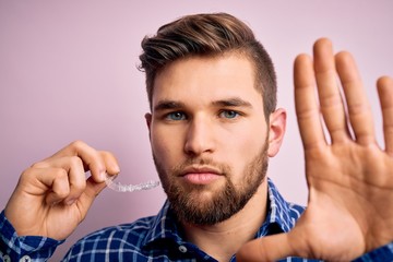 Young blond man with beard and blue eyes holding dental aligner over pink background with open hand...