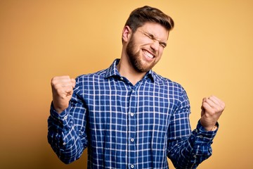 Young blond businessman with beard and blue eyes wearing shirt over yellow background very happy and excited doing winner gesture with arms raised, smiling and screaming for success. Celebration
