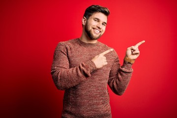 Young blond man with beard and blue eyes wearing casual sweater over red background smiling and looking at the camera pointing with two hands and fingers to the side.