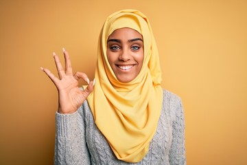 Young beautiful african american girl wearing muslim hijab over isolated yellow background smiling positive doing ok sign with hand and fingers. Successful expression.