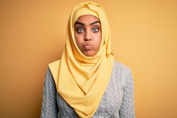 Young beautiful african american girl wearing muslim hijab over isolated yellow background puffing cheeks with funny face. Mouth inflated with air, crazy expression.