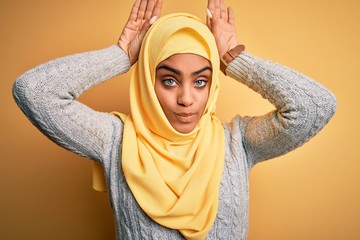 Young beautiful african american girl wearing muslim hijab over isolated yellow background Doing bunny ears gesture with hands palms looking cynical and skeptical. Easter rabbit concept.