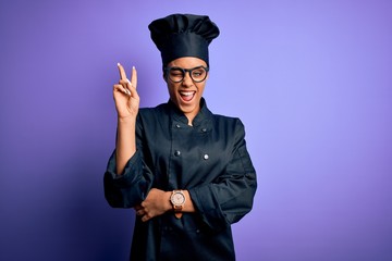 Young african american chef girl wearing cooker uniform and hat over purple background smiling with happy face winking at the camera doing victory sign with fingers. Number two.