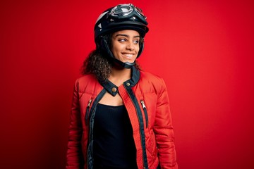 Young african american motorcyclist girl wearing moto helmet and glasses over red background looking away to side with smile on face, natural expression. Laughing confident.