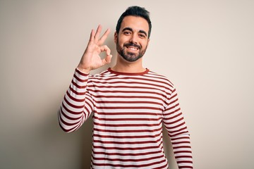 Young handsome man with beard wearing casual striped t-shirt standing over white background smiling positive doing ok sign with hand and fingers. Successful expression.