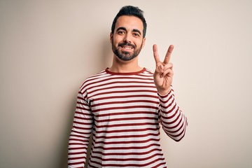 Young handsome man with beard wearing casual striped t-shirt standing over white background showing and pointing up with fingers number two while smiling confident and happy.