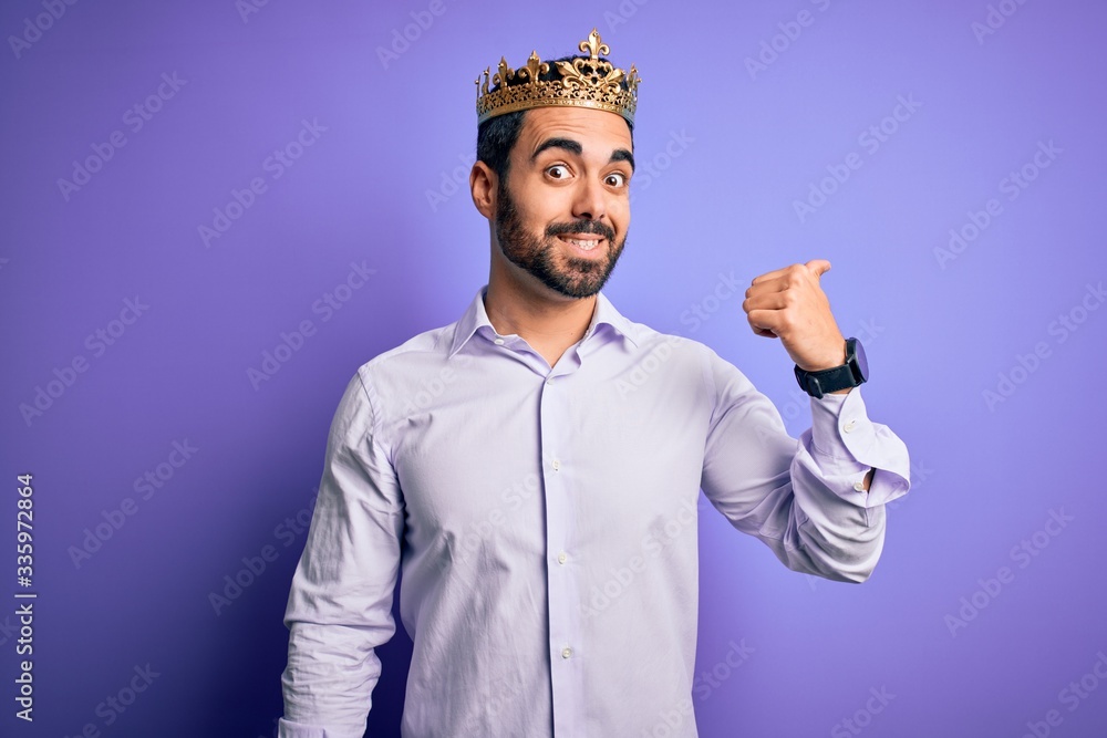 Sticker Young handsome man with beard wearing golden crown of king over purple background smiling with happy face looking and pointing to the side with thumb up.