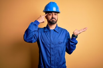 Mechanic man with beard wearing blue uniform and safety helmet over yellow background confused and annoyed with open palm showing copy space and pointing finger to forehead. Think about it.