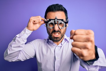 Young handsome optical man with beard wearing optometry glasses over purple background annoyed and frustrated shouting with anger, crazy and yelling with raised hand, anger concept