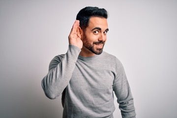 Young handsome man with beard wearing casual sweater standing over white background smiling with hand over ear listening an hearing to rumor or gossip. Deafness concept.