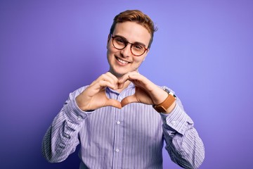 Young handsome redhead man wearing casual shirt and glasses over purple background smiling in love doing heart symbol shape with hands. Romantic concept.