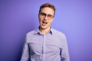 Young handsome redhead man wearing casual shirt and glasses over purple background winking looking at the camera with sexy expression, cheerful and happy face.