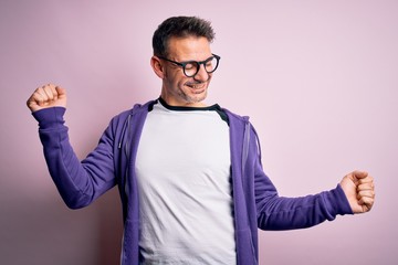 Young handsome man wearing purple sweatshirt and glasses standing over pink background Dancing happy and cheerful, smiling moving casual and confident listening to music