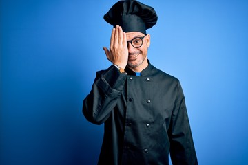 Young handsome chef man wearing cooker uniform and hat over isolated blue background covering one eye with hand, confident smile on face and surprise emotion.